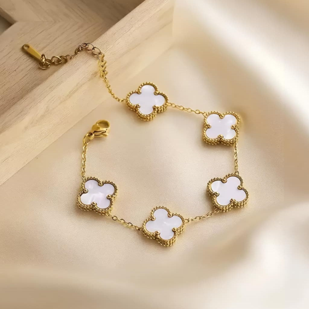 A gold-tone chain bracelet with five white clover-shaped links, styled on a beige satin fabric next to a light wood tray.