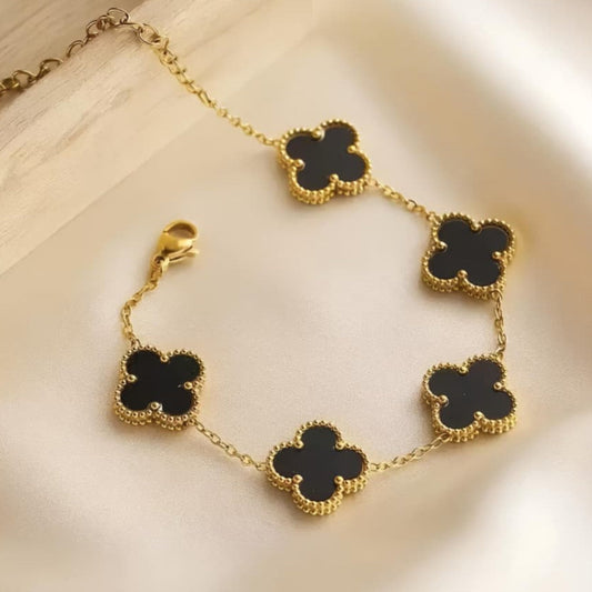 A gold chain bracelet with five black clover-shaped motifs, styled on a soft cream satin fabric next to a light wood block.