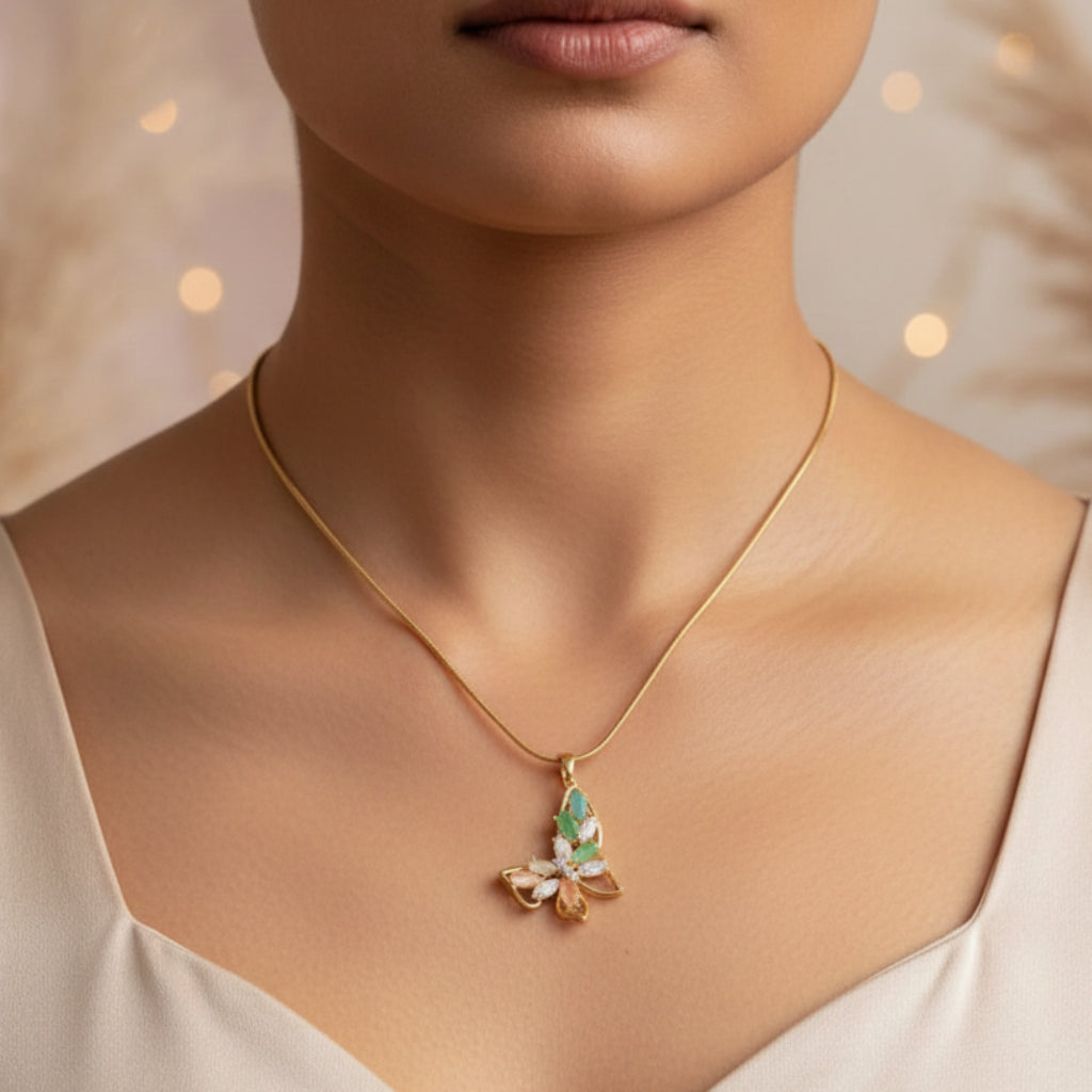 A gold chain necklace with a butterfly pendant featuring green and pink iridescent details, displayed on a cream-colored jewelry bust against a warm, softly blurred background with bokeh lights.